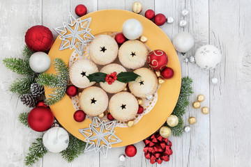 Christmas mince pies on a gold plate with holly, fir, red and white bauble decorations with foil wrapped chocolates on rustic wood background.  