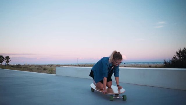 Clumsy cute teenage girl or young woman sits on knees on top of longboard, can not stand up, learns how to ride skateboard, laughs and makes funny faces, enjoying herself on boardwalk