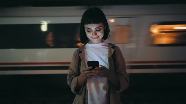 Beautiful young woman in trendy coat stands on platform in evening, smartphone lights up her face, she texts on messaging application, smiles and giggles, train passes by