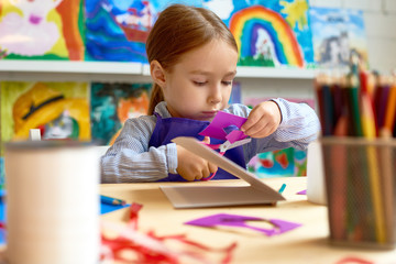 Portrait of cute little girl carefully cutting out shapes while making collage during art class in...