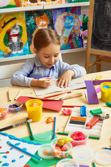 Portrait of adorable little girl writing wishes on handmade gift card sitting at messy  table with supplies during art and craft class of pre-school