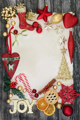Decorations and symbols of christmas forming a border with gold joy sign, baubles, holly, mistletoe, fir, mince pie, cinnamon sticks and dried orange on rustic wood background.