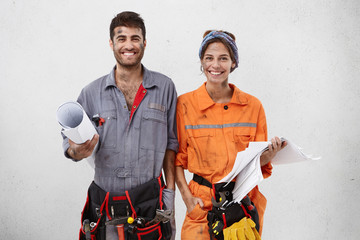 Joyful handyman and female architect collaborate together, wear working clothes, hold sketches with...