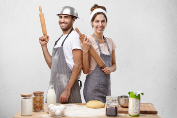 Positive participants of culinary show stand back to each other, keep rolling pins, smile pleasantly as being glad to be popular on tv. Cookers rivals take part in competition at kitchen, show talents