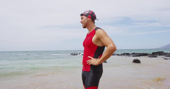 Triathlon swimming - male triathlete swimmer standing on beach after swim training. Fit man resting after swimming freestyle crawl in professional triathlon suit while training for ironman on Hawaii.