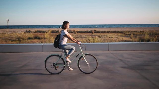 Young petite woman in white tshirt and denum jeans with backpack rides her bicycle next to beach and sunset over sea, she pedals through promenade boardwalk,enjoys sun