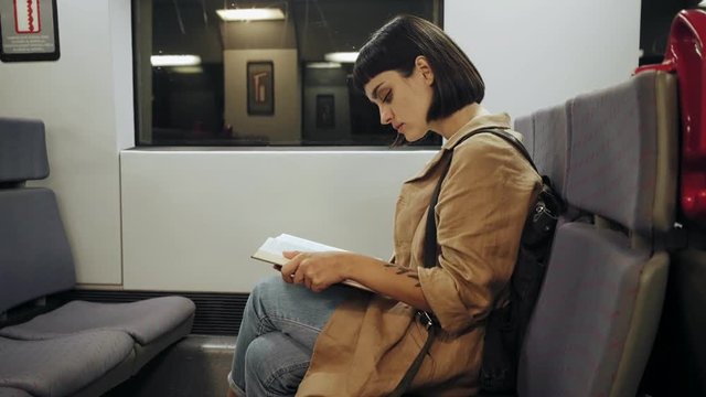 Pretty cute young woman in hipster coat sits in midnight express train on her way back home in commuting hours, she reads book, looks out of window on landscapes passing by
