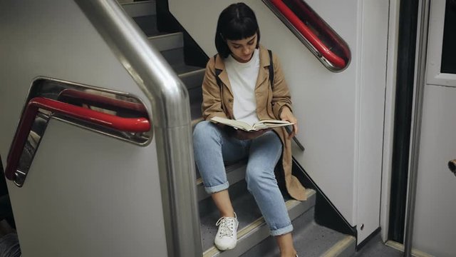 Top view on attractive brunette student girl is reading a book sitting on stairs of double decker train car during her commute trip