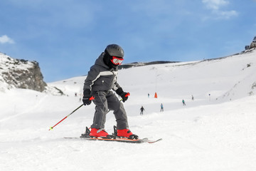 A little brave toddler boy in full ski equipment races downhill with blue sky on the background at winter ski resort