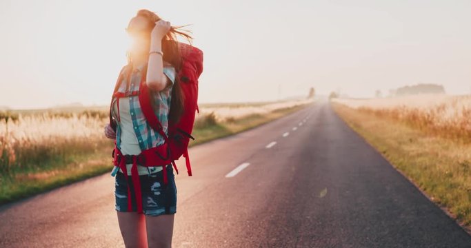 Traveler woman walking on a sunny road. Young happy backpacker woman starting a journey on a sunlit country road. Slow motion, 120fps. 4K, DCi. Poland, Lower Silesia