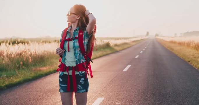 Traveler woman walking on a sunny road. Young happy backpacker woman starting a journey on a sunlit country road. Slow motion, 120fps. 4K, DCi. Poland, Lower Silesia