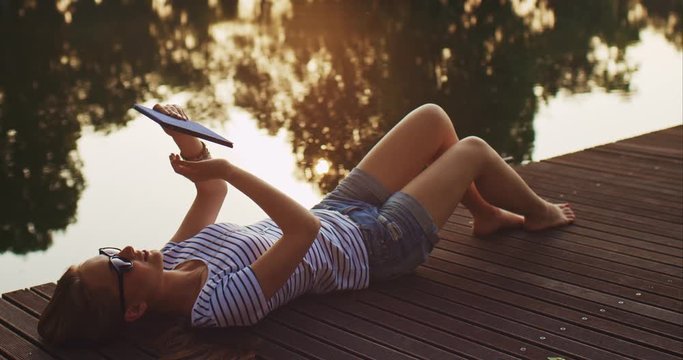 Woman using digital tablet outdoors, relaxing on a wooden jetty near the lake with sunny background, lens flare.  slow motion. 