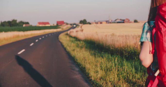 Traveler woman hitchhiking on a sunny road and walking. Young happy backpacker woman looking for a ride to start a journey on a sunlit country road. Slow motion, 120fps. 4K, DCi. Poland, Lower Silesia