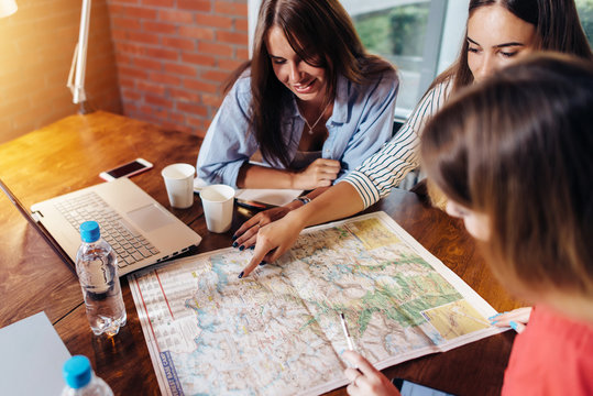 Smiling Female Friends Sitting At Desk Planning Their Vacation Looking For Destinations On Map