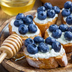 Crostini with ricotta cheese, blueberries and honey on a wooden plate, square format