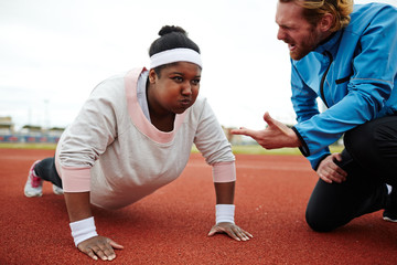 Dedicated sport trainer encouraging overweight young woman try harder while practicing push-ups