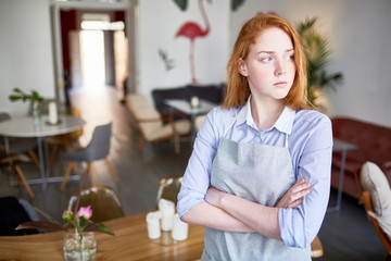 Serious waitress in uniform crossing her arms on chest and looking through window of cafe