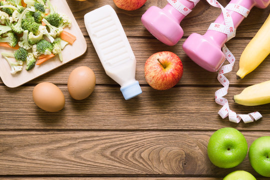 Healthy eating, dieting, slimming and weight loss concept - Top view of green apple, bananas, measuring tape, dumbbells, eggs, milk bottle and vegetables block Kerry and carrot on wooden background.