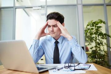 Tired young financial manager suffering from terrible headache while preparing annual accounts with help of laptop, interior of spacious office on background