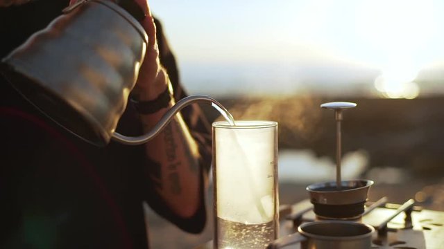 guy in black t-shirt with tattoo on his arm pouring hot water from kettle to glass bowl on seashore slow motion 