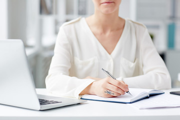 Unrecognizable young businesswoman wearing white shirt sitting at office desk and taking necessary notes, blurred background