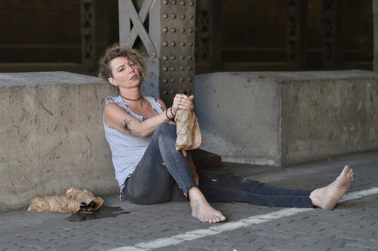 a punk woman under a bridge with bottles of alcohol nearby and a bottle in a brown paper bag in her hand 