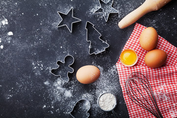 Cookies cutters, flour, rolling pin, eggs and whisk on kitchen table top view. Christmas baking background.
