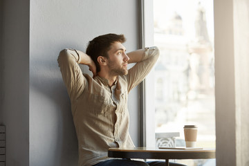 Attractive young unshaven guy sitting in cafe, drinking coffee, looking at window with hands behind...