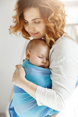 Portrait of young mother and newborn son sleeping on mother chest in blue baby sling. Family happiness vibes. Family concept.