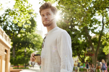 Young handsome guy with ginger hair and beard in white shirt drinking coffee and waiting his girlfriend for a date in city garden.