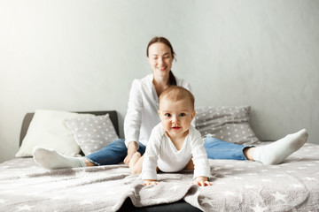 Portrait of sweet little baby sitting with mother on bed and crawling in camera direction with interested and excited expression.