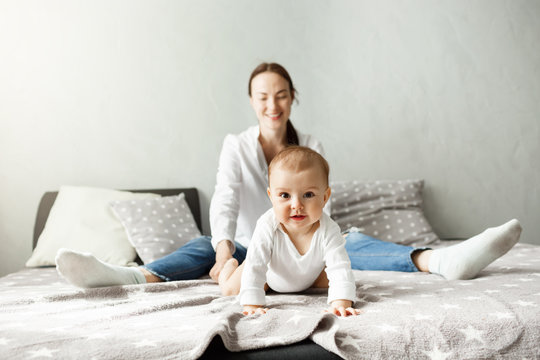 Portrait of sweet little baby sitting with mother on bed and crawling in camera direction with interested and excited expression.