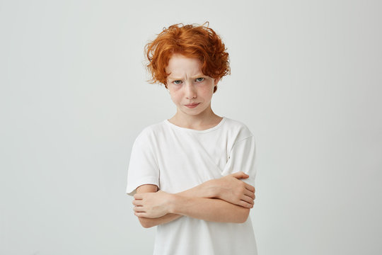 Portrait of unhappy red haired boy with freckles looking in camera with upset expression, crossing hands being dissatisfied that his mom scolded him.