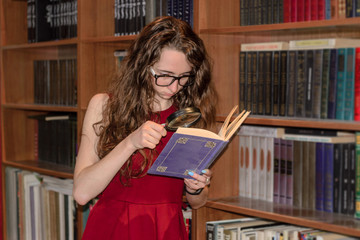 Female student in glasses is looking through the magnifying glass in the book.  Library in background