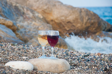 Two wineglasses with red, white drinks stay on grey stone on sea beach
