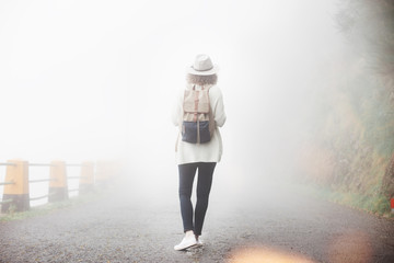 Young traveler girl wearing backpack, sneakers and hat standing in fog in forest on the road.