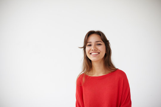 Picture of attractive cheerful young Hispanic lady dressed in oversize red sweater grinning broadly at camera, her eyes expressing joy. Positive friendly looking girl posing isolated in studio