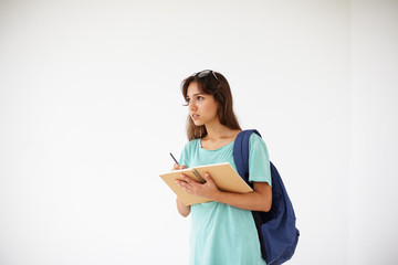 Thoughtful dreamy female student wearing casual blue shirt and glasses on head making notes in her personal diary. Cute pensive student girl with backpack doing homework, writing composition or essay