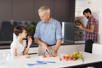 The family is preparing a salad for Thanksgiving. The old man cuts vegetables, the boy eats an apple