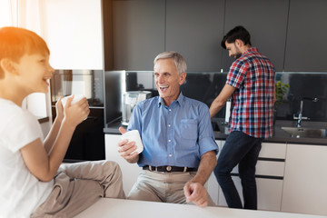 The old man and the boy laugh at something while the boy's father washes the dishes behind them