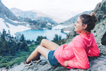 Young woman sitting and resting high in mountains. Risky rock climbing in peaceful wilderness area. Enjoying amazing snowy lake view
