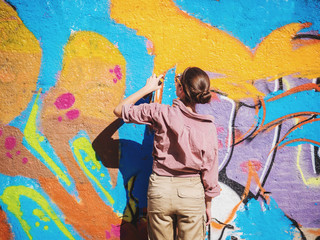 Naklejka premium Piękna młoda dziewczyna robi kolorowemu graffiti z aerozolową kiścią na miastowej ulicy ścianie. Filmowy stonowany strzał. Kreatywna sztuka. Utalentowana kobieta w szkłach rysuje obrazek.