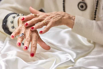 Foto auf Alu-Dibond Senior manicured hands with jewelry. Old aristocratic woman hands with beautiful red manicure wearing luxury rings, white silk. Female treatment and wealth. © DenisProduction.com