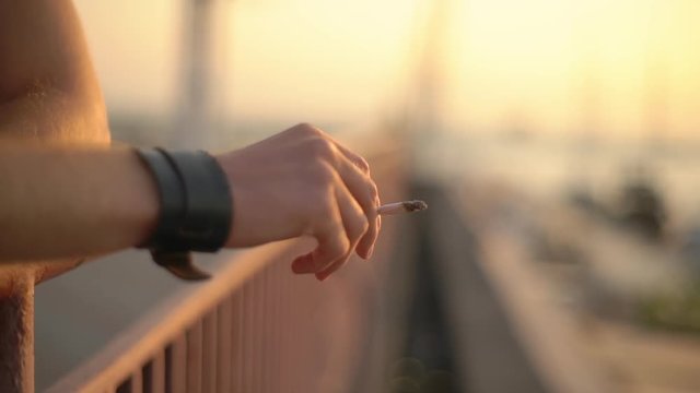 Male hand with black whistband holding a rollup cigarette, ash falling. Bridge view, sunset. Slow motion.