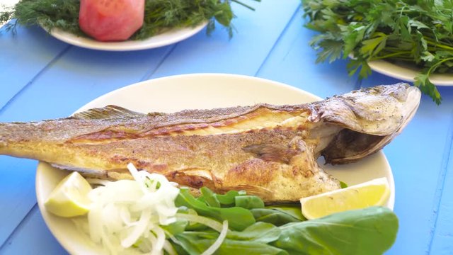 Grilled sea bass on plate with onions, sliced lemon and garden rocket aragula on blue table. Greek food.