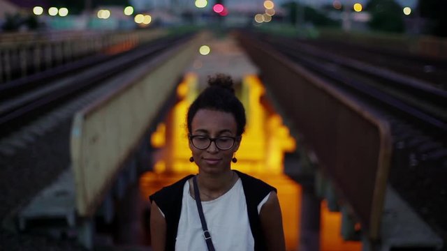 Cute black girl looks straight at camera, open smile, white shirt. Urban evening background, cars passing by, bridge. Slow motion.