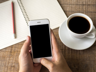 Close up business man's hand holds smart phone with black isolated screen over background of notebook, coffee on wooden background