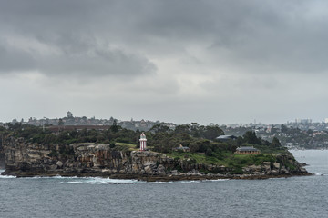 Fototapeta na wymiar Sydney, Australia - March 21, 2017: South Head cliffs and park with short Hornby lighthouse, backed by green vegetation under foggy dark cloudscape. Part of city skyline in distance.