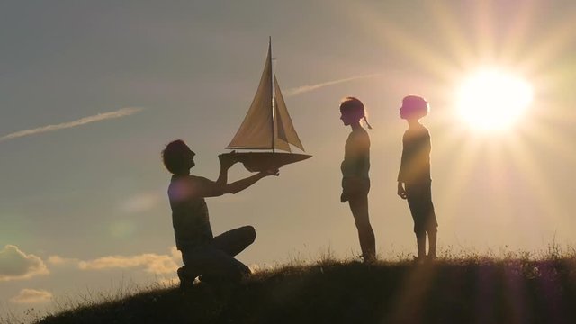 A model of a sailboat. A man is holding a ship model against the sky. The father is giving the ship to the child. Backlight. Silhouettes of people against the sky and the sun.