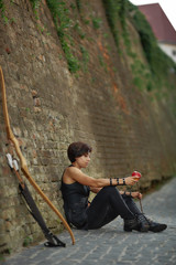 Woman leaning on brick wall near bow and arrow.